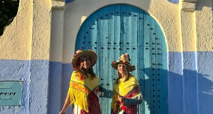 Deux femmes portant de brillants chapeaux traditionnels et des châles posent devant une porte voûtée bleue à Chefchaouen.