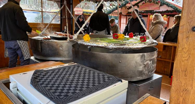 Étal de restauration avec de grandes poêles de grill rondes cuisant des saucisses et des légumes tandis que les clients attendent à proximité.