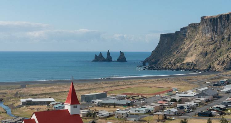 Un village côtier islandais avec une église aux toits rouges surplombe les formations rocheuses de Reynisdrangar au-delà des plages de sable noir.