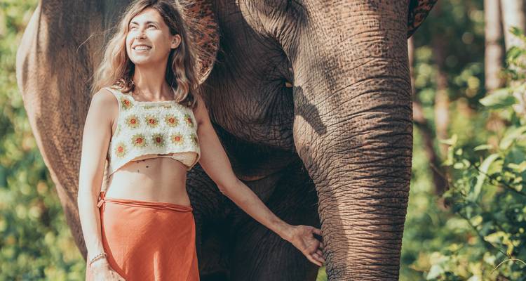 A cheerful woman stands beside an elephant, sunlight filtering through green foliage behind.