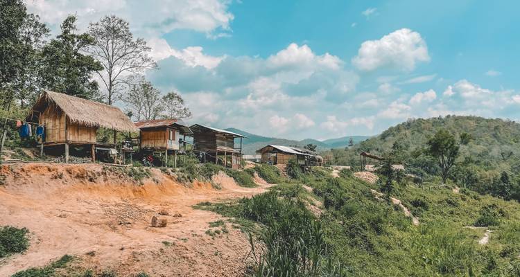 Simple wooden huts perched on an orange-soil ridge overlooking green hills and blue sky.