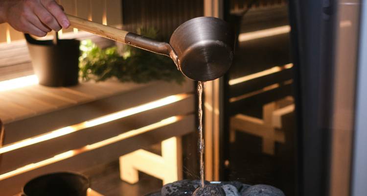 Una mano vierte agua de un cucharón de madera sobre las piedras calientes de la sauna, enviando vapor a una habitación de cedro iluminada calurosamente.