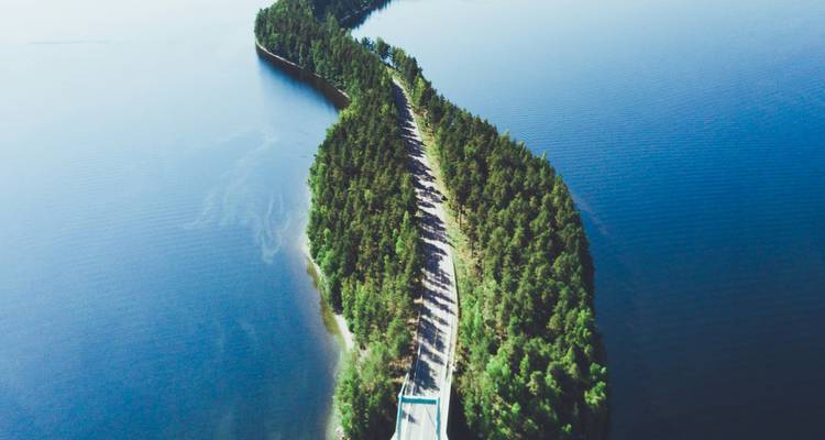 Una vista aérea de un istmo estrecho y boscoso con una carretera que atraviesa las aguas azul profundo de un lago en la región de lagos de Finlandia.