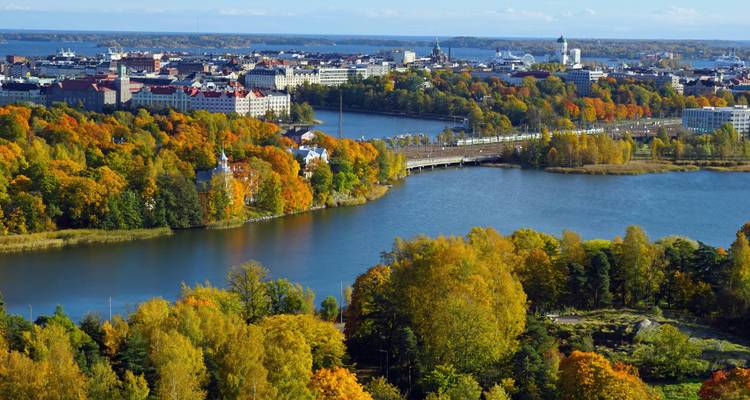 Un colorido panorama otoñal de Helsinki con follaje naranja enmarcando las vías fluviales y el horizonte de la ciudad bajo un cielo brillante.