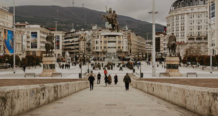 La plaza central de Skopie con una gran estatua ecuestre de Alejandro Magno, rodeada de edificios de mármol y colinas distantes.