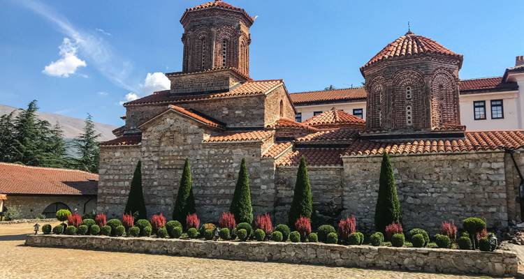 Monasterio histórico de piedra con tejados de tejas rojas rodeado de arbustos y flores bien cuidados bajo un cielo despejado cerca del Lago Ohrid.