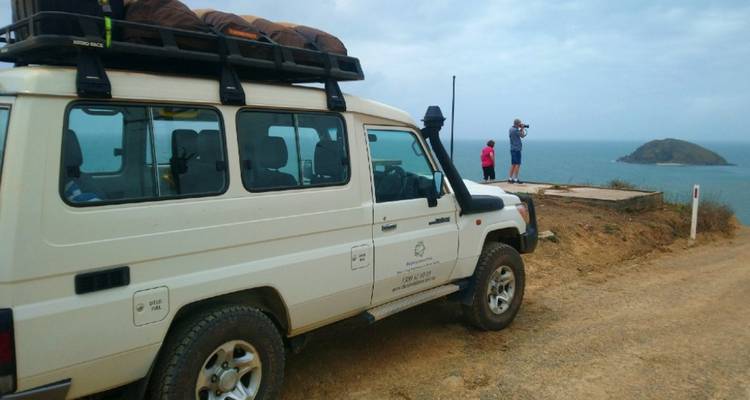 Un véhicule tout-terrain 4x4 garé sur un sentier côtier accidenté tandis que deux voyageurs photographient la vue océanique lors d'une journée nuageuse.
