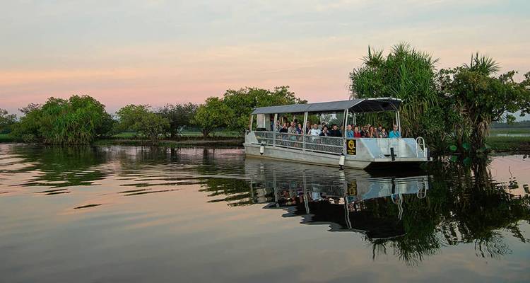 Rondvaartboot zonder zijwanden vol passagiers glijdt over rustig moeraswater dat de zachte zonsonderganglucht en weelderige oeverbegroeiing weerspiegelt.