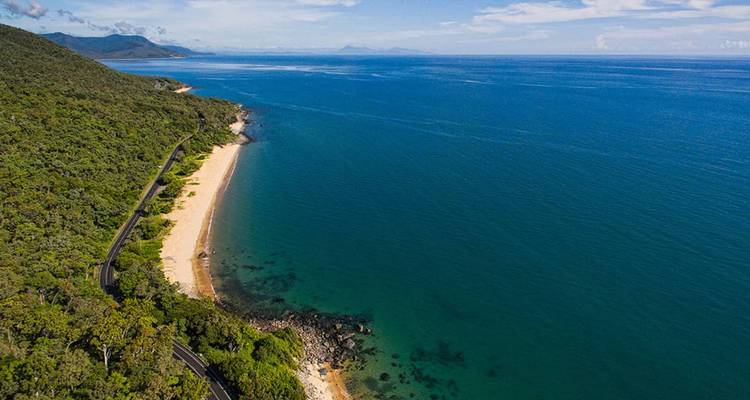 Vue aérienne d'une route traversant une forêt tropicale côtière qui épouse une courbe de mer turquoise éclatante et de sable doré.