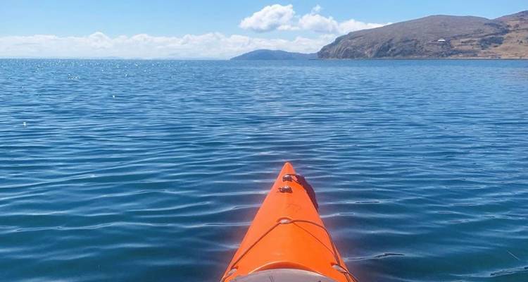 Proue d'un kayak orange tranchant les eaux bleu profond calme d'un lac en direction des montagnes lointaines sous des ciels dégagés