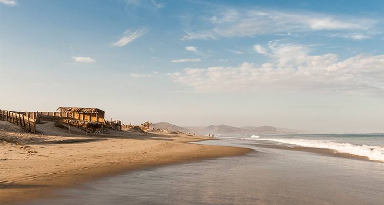 Une plage de sable tranquille s'étend le long de la côte avec une cabane en bois rustique et des vagues douces sous des cieux bleus.