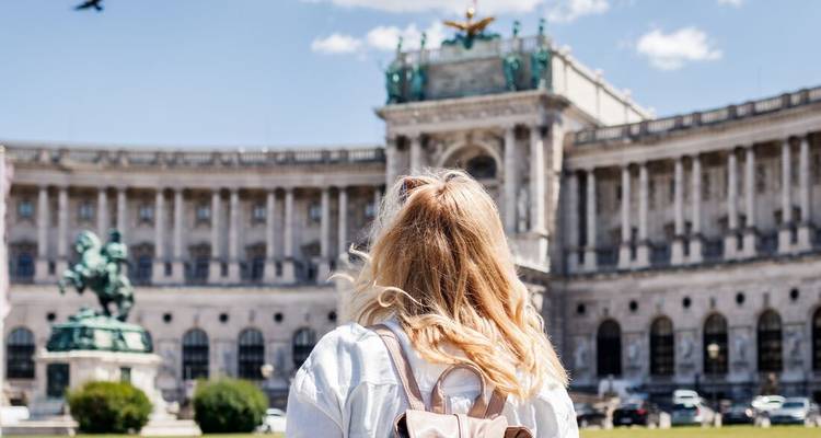 Vista trasera de una viajera rubia con mochila admirando el grandioso Palacio de Hofburg en Viena bajo un cielo azul.