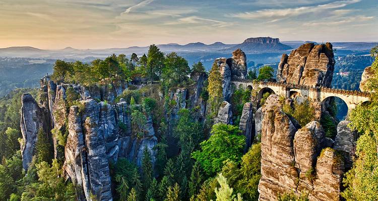 Vista aérea del dramático Puente Bastei que se extiende sobre imponentes pilares de arenisca en un paisaje boscoso al amanecer.