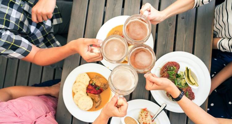 Des amis trinquent avec de grandes chopes de bière au-dessus de plats copieux d'Europe centrale sur une table de terrasse en bois.