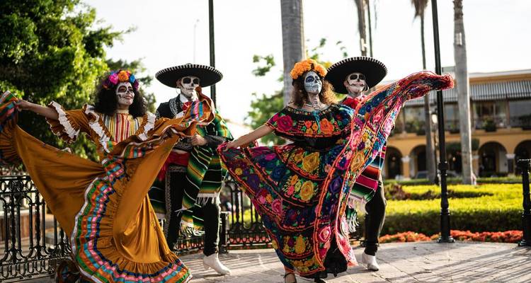 Artistas disfrazados con maquillaje del Día de los Muertos giran vestidos tradicionales coloridos en una plaza.