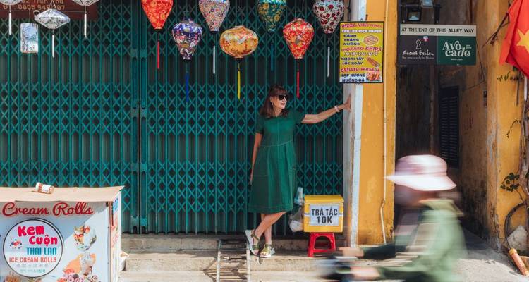 Una mujer con un vestido verde posa junto a faroles colgantes de colores fuera de una tienda con persianas cerradas mientras una scooter pasa a toda velocidad.