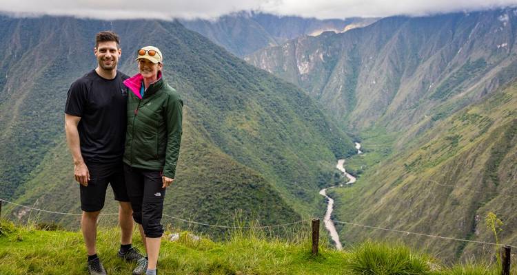 Lächelndes Paar posiert auf einem grasbewachsenen Kamm mit Blick auf ein tiefgrünes Andental mit einem sich windenden Fluss darunter.