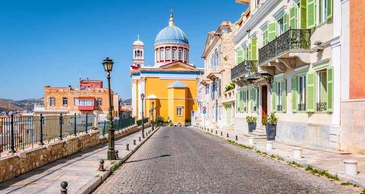 Rue néoclassique colorée à Syros avec façade d'église éclatante et dôme bleu sous un ciel dégagé.