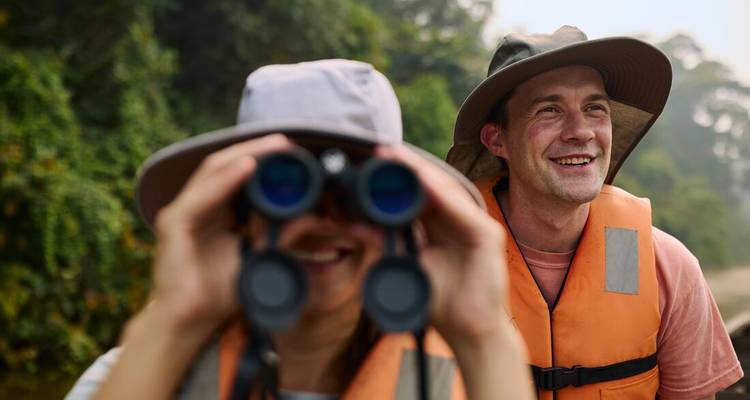 Deux personnes portant des chapeaux et des gilets de sauvetage, avec des jumelles dans la forêt.