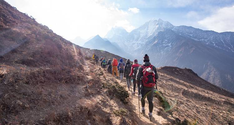 Een lange rij wandelaars klimt over een stoffig Himalayaans pad omhoog naar torenhoge, sneeuwbedekte pieken in het felle zonlicht.