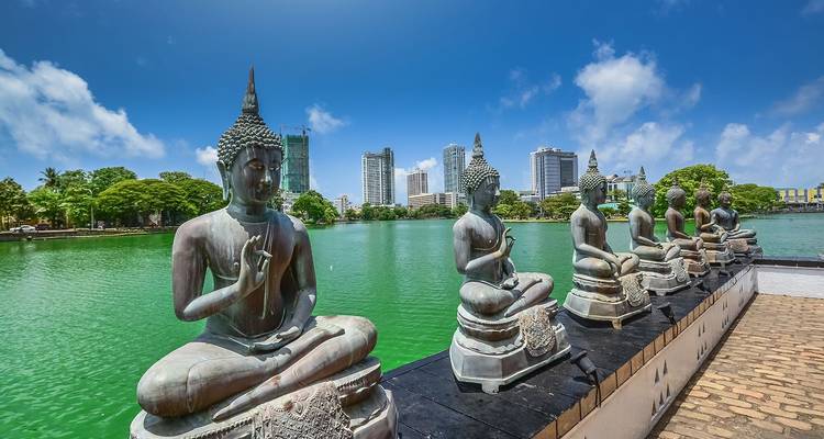 Rangée de statues de Bouddha assis à côté d'un lac d'émeraude avec les gratte-ciel modernes de Colombo en arrière-plan.