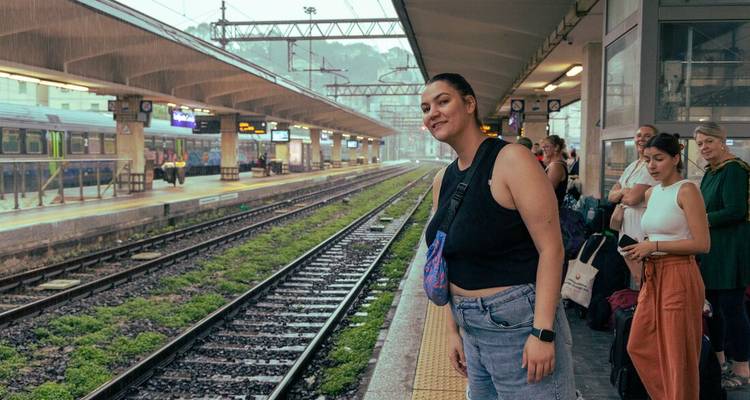 Des voyageurs attendent sur un quai de gare pluvieux tandis qu'une femme pose près des voies.