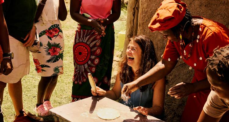 Reizigers lachen terwijl ze leren plat brood te bereiden met lokale vrouwen buiten in een Maasai-dorp.