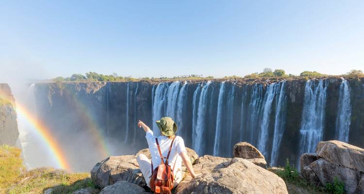 Een reiziger zit op rotsen aan de rand van een klif en wijst naar de Victoria Falls waar een regenboog door de mist boogvormig loopt.