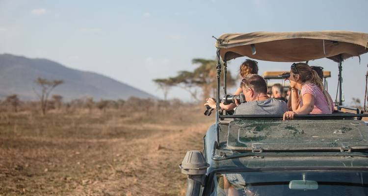 Toeristen die door het dakluik van een 4x4 safariauto omhoog staan en wilde dieren fotograferen op een open savanne met heuvels in de verte.
