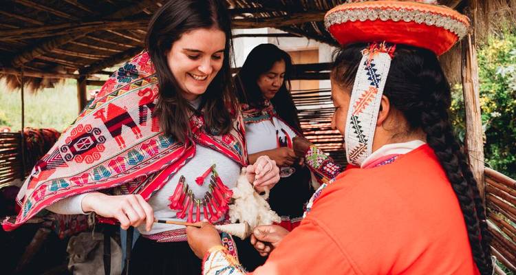 Een reiziger leert traditioneel spinnen van een Andesvrouw in levendige geborduurde kleding in een werkplaats met rieten dak.