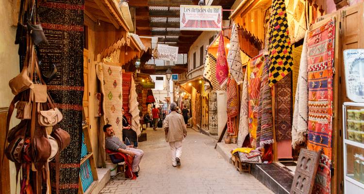 Bustling Moroccan souk lined with vivid carpets and leather goods, shoppers and locals in the aisle