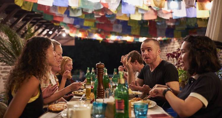 Una cena al aire libre llena de vida bajo filas de banderas de oración ondulantes, con viajeros compartiendo historias.