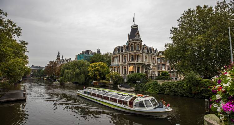 Un bateau-promenade à toit de verre navigue devant d'élégantes demeures et des arbres feuillus le long d'un canal d'Amsterdam.
