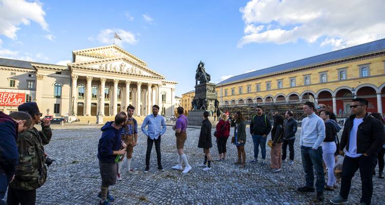 Un groupe de touristes se rassemble sur une spacieuse place pavée face au Théâtre national néoclassique de Munich.