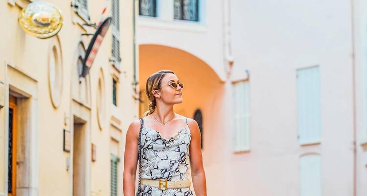 Une femme élégante se promène dans une ruelle étroite aux couleurs pastel, portant des lunettes de soleil et une robe à motifs.