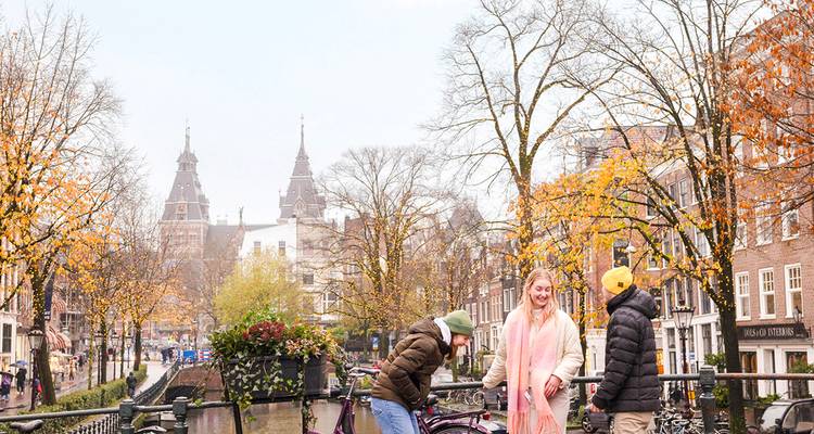 Des amis profitent d'une journée d'automne sur un pont de canal d'Amsterdam bordé de vélos et d'arbres dorés.