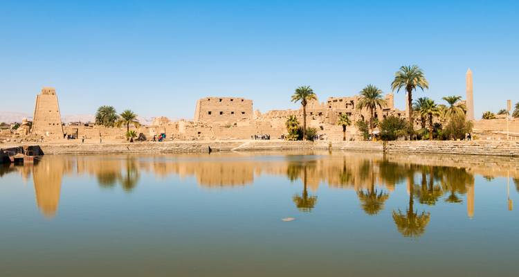 Heilig meer bij Karnak Tempel weerspiegelt zandstenen ruïnes, obelisk en palmbomen onder een heldere blauwe lucht.