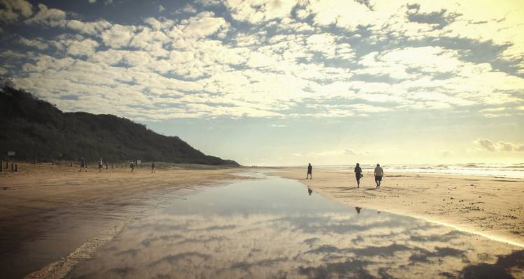 Menschen, die an einem Strand spazieren gehen, mit dramatischen Wolken, die sich im Wasser spiegeln.