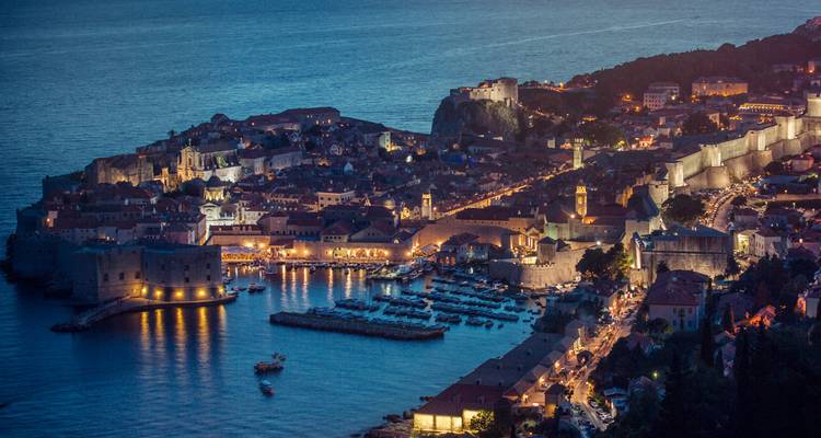 Vue aérienne au crépuscule de la ville fortifiée illuminée de Dubrovnik et de son port entourés par une mer d'un bleu profond.