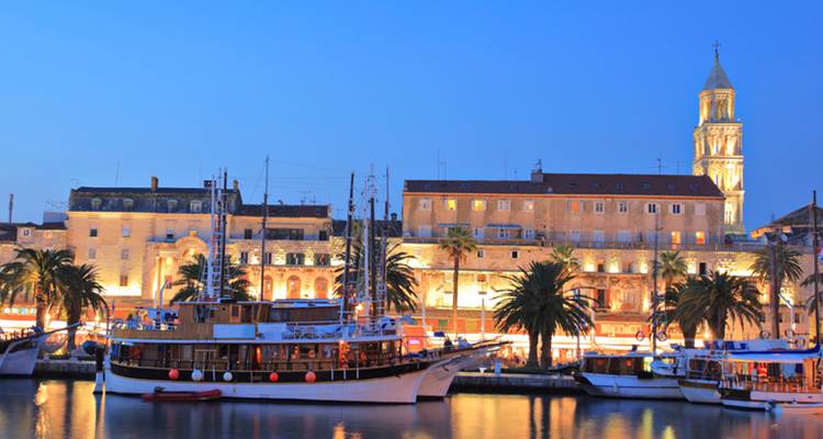 Les lumières du soir le long du front de mer de Split avec des bateaux ancrés dans des reflets calmes et le clocher illuminé.