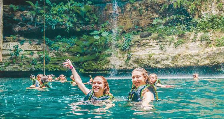 Travellers swim and splash in the clear blue waters of a lush Mexican cenote with a small waterfall.