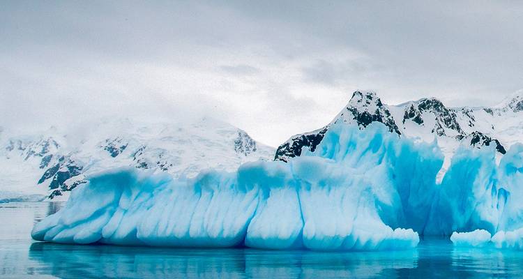 Iceberg bleu sculpté avec des montagnes enneigées s'estompant dans un ciel nuageux