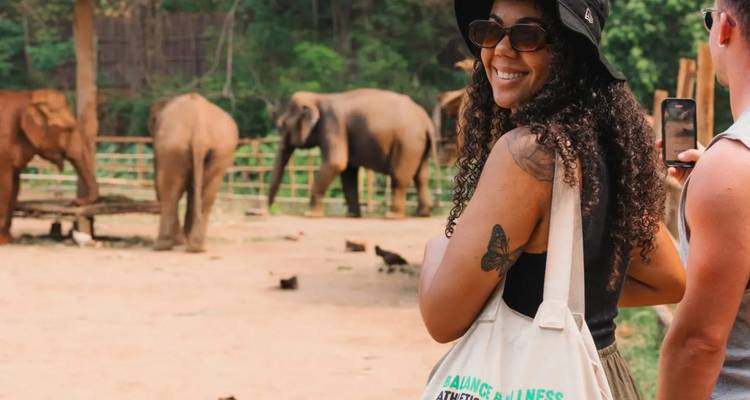 Smiling visitor with tote bag observes elephants in a sanctuary enclosure.