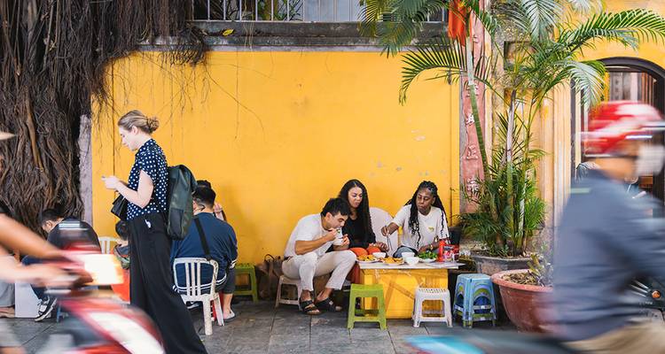 Los lugareños y visitantes disfrutan de comida callejera en taburetes bajos junto a una pared amarillo brillante mientras las motocicletas pasan borrosas.