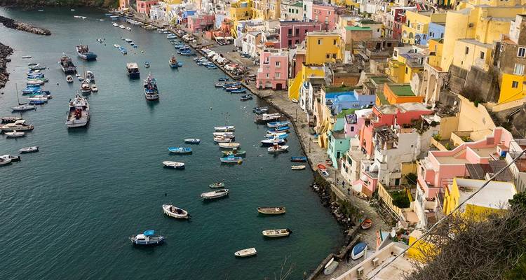 Des maisons aux couleurs pastel vibrantes se regroupent le long du port de Procida avec des bateaux de pêche parsemant l'eau turquoise.