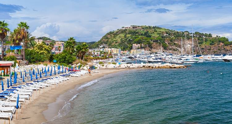 Plage de sable incurvée bordée de parasols bleus et blancs face à une baie calme adossée à des collines vertes.