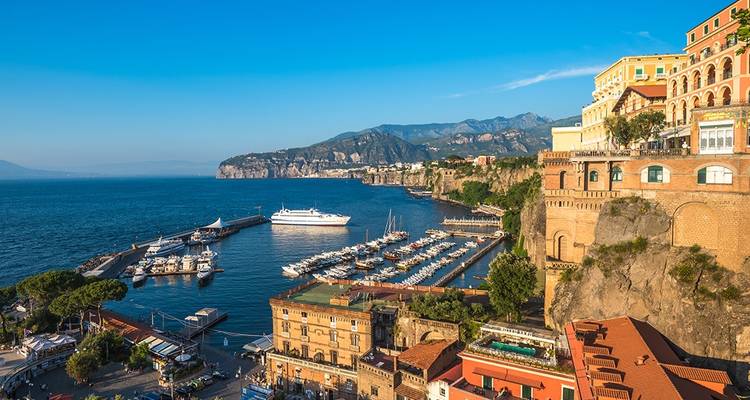 Vue panoramique sur la marina de Sorrente, les falaises en terrasses et les montagnes lointaines baignées de lumière dorée.