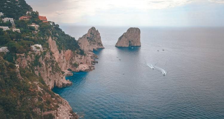 Piles de Faraglioni au large de Capri vues depuis une haute falaise sous une lumière d'après-midi brumeuse avec des sillages de bateaux en contrebas.