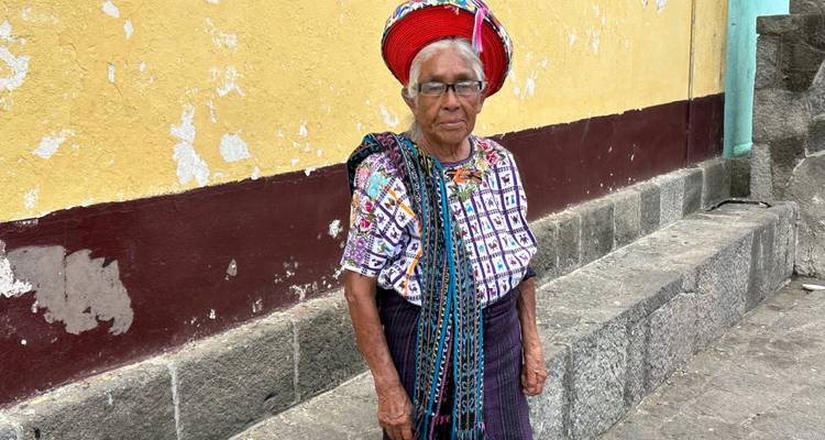 Elderly woman in traditional clothing standing against a wall.