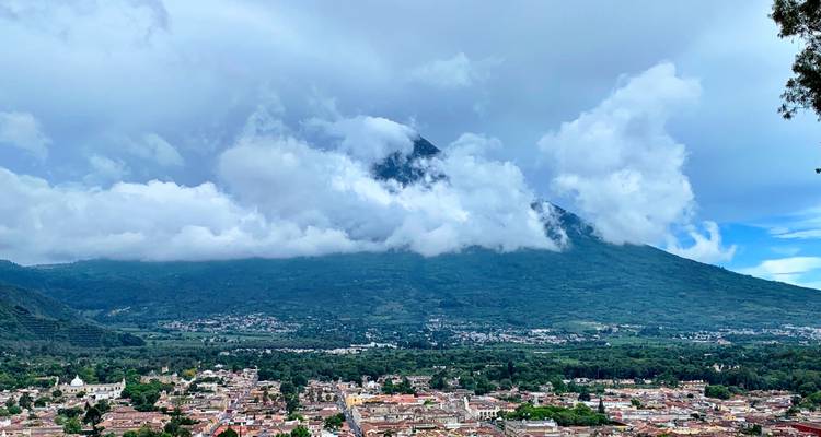 A panoramic view of a city with a volcano in the background, partially covered by clouds.
